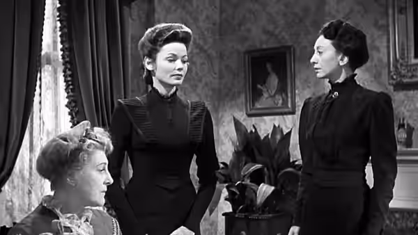 Three women gather in a furnished room, two standing beside a seated woman