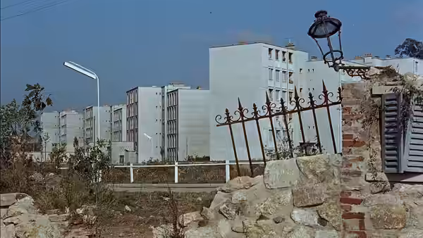 A modern white house rises behind a stone wall and ornate iron gate
