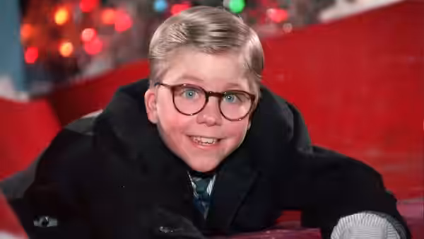 Ralphie (Peter Billingsley) smiles while bundled in a heavy coat beside Christmas lights