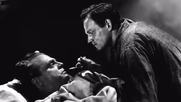 Cody Jarrett (James Cagney) leans over a man lying on a bed