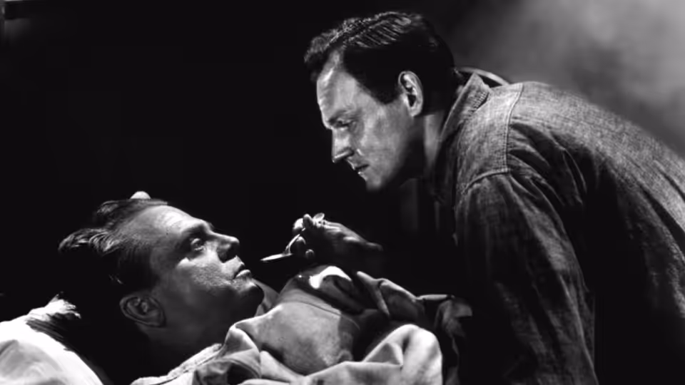 Cody Jarrett (James Cagney) leans over a man lying on a bed