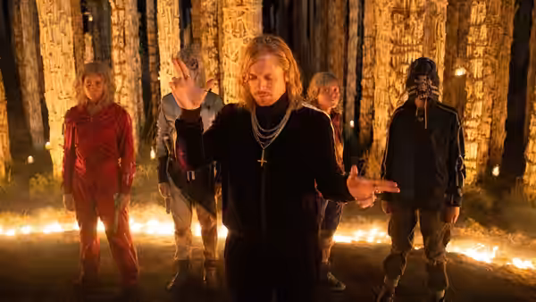 Sir Jimmy Crystal (Jack O'Connell) gesturing with arms outstretched among followers in a forest