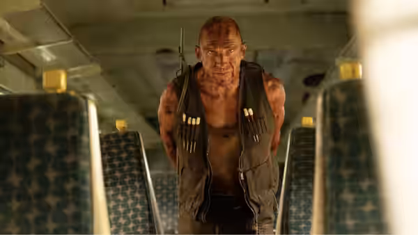 a muscular bald man with ammunition vest crouching inside a vehicle