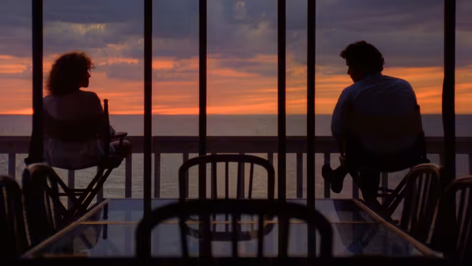 a man and a woman seated in director's chairs on a balcony overlooking the ocean at sunset