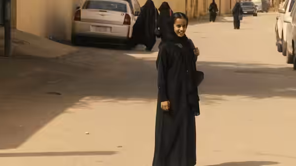 Young girl in black abaya standing on a sunlit street with cars parked nearby