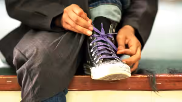 Hands tying purple laces on a worn black sneaker resting on a wooden bench