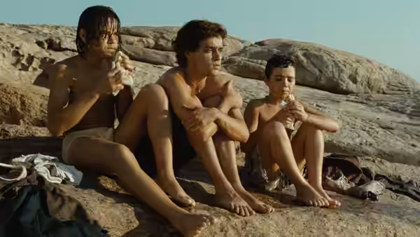Three shirtless boys sit on rocks by the sea eating snacks