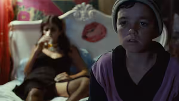 A boy in a beanie stands in front of a woman drinking on a bed