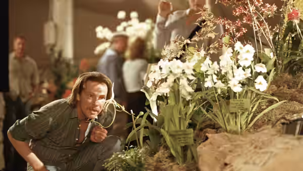 John Laroche (Chris Cooper) crouching beside white orchids at a display