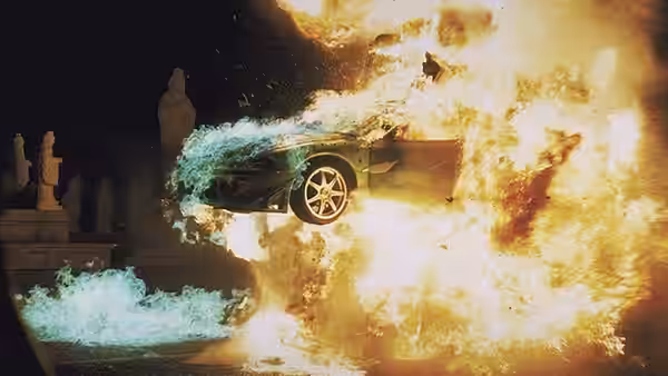 A car erupts in flames beside statues as debris scatters through the air