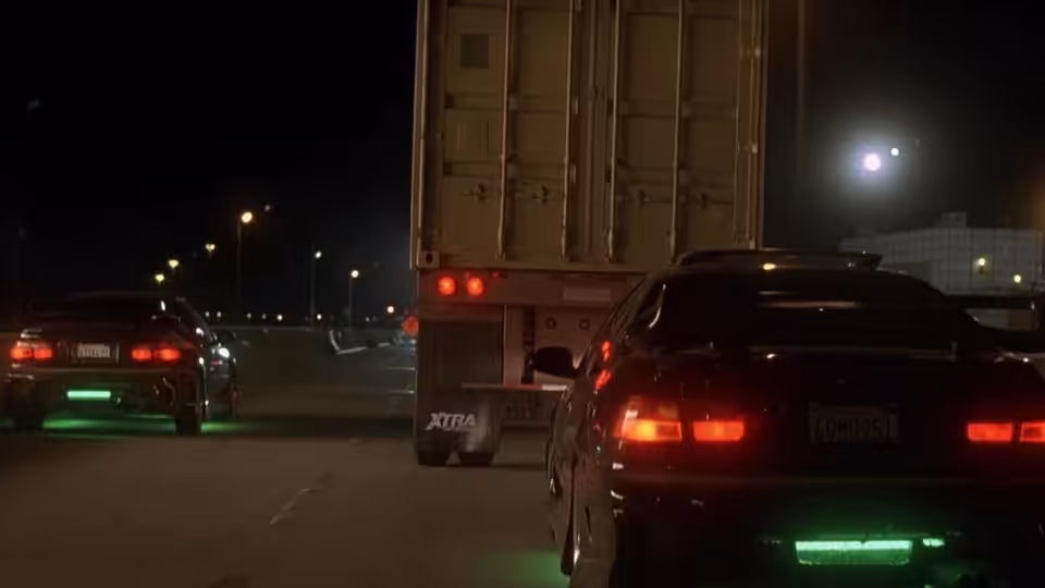 Two cars speed after a truck along a dark highway at night