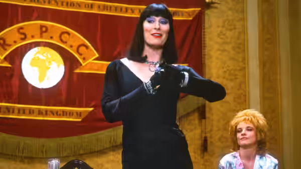 Miss Eva Ernst (Anjelica Huston) stands beside a blonde woman beneath a red banner