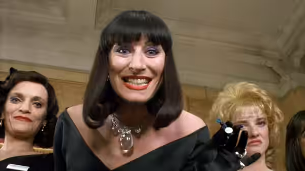 Miss Eva Ernst (Anjelica Huston) grins and holds up a small object among other women