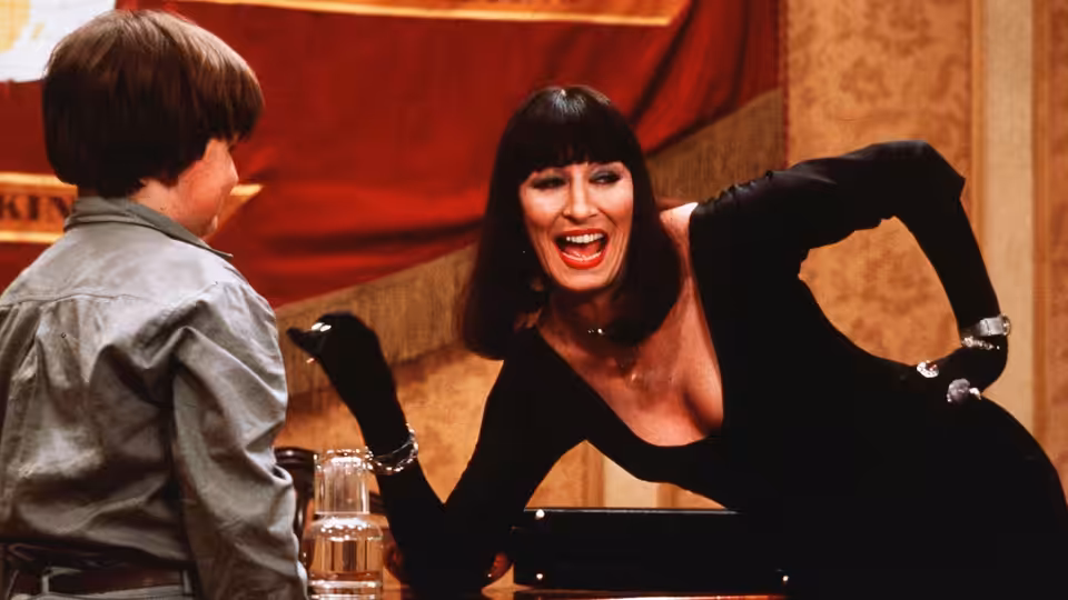 Miss Eva Ernst (Anjelica Huston) leans across a table toward a boy in a hotel room