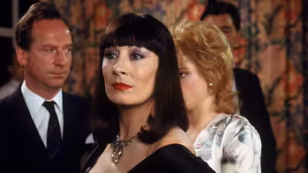 Miss Eva Ernst (Anjelica Huston) stands among formally dressed guests in a hotel room