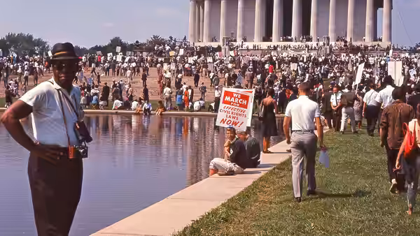 Man with camera stands by reflecting pool as large crowd gathers at Lincoln Memorial