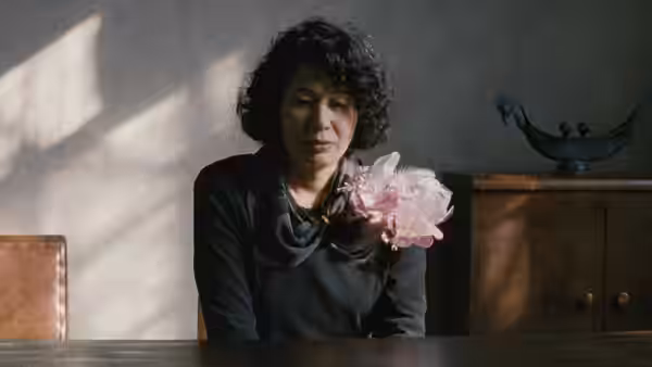 A woman holds a pink flower while seated at a table