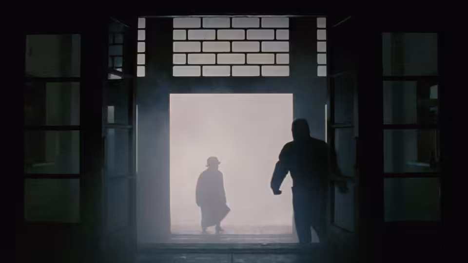 Two figures meet at a bright doorway inside a shadowy building