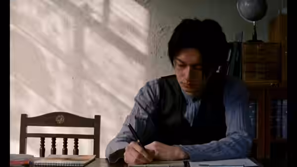A man writes in a notebook at a desk in a sunlit room