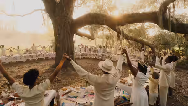 A crowd joins hands around a large tree at an outdoor gathering