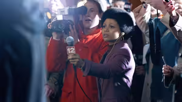 Linda Page (Myha'la Herrold) holding a microphone while surrounded by reporters and cameras