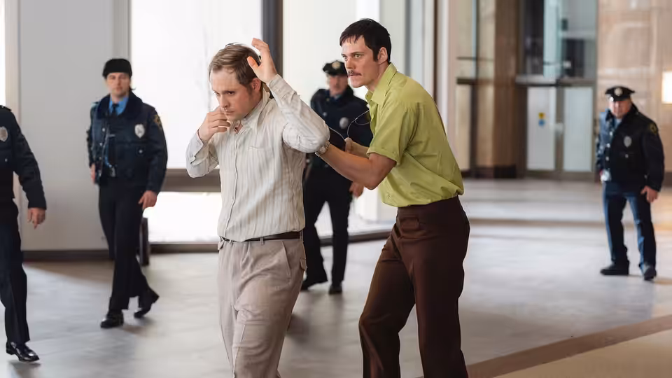 Tony Kiritsis (Bill Skarsgård) held by Richard Hall (Dacre Montgomery) inside a lobby with police