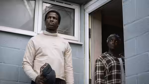 Bol Majur (Sope Dirisu) stands outside a doorway as Rial watches from inside