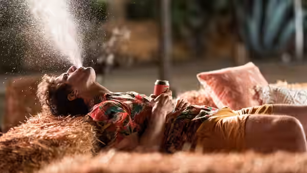 Nyles (Andy Samberg) lies on a hay-covered lawn holding a drink as water sprays overhead