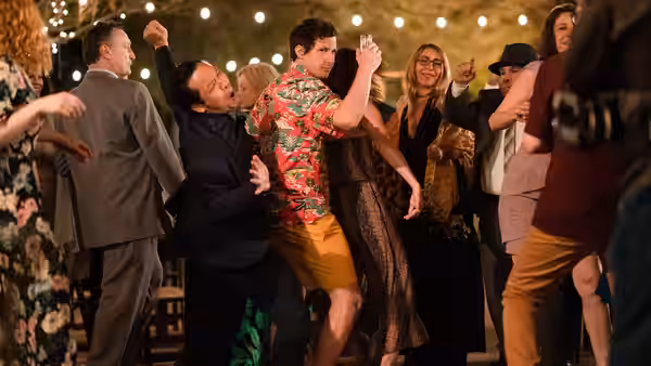 Nyles (Andy Samberg) dances with a drink among guests at a string-lit wedding reception
