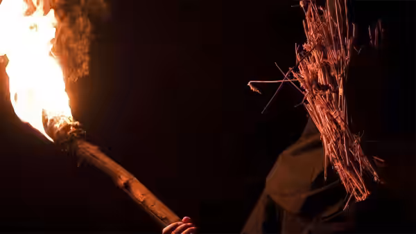 Figure holds flaming torch beside masked companion in darkness