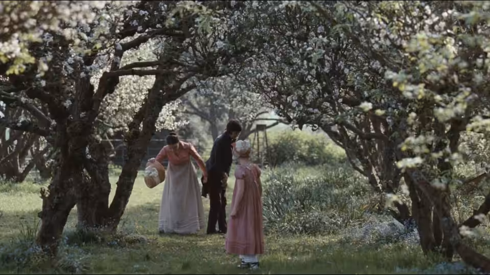 Two women stand beneath flowering trees in an orchard, one carrying a basket