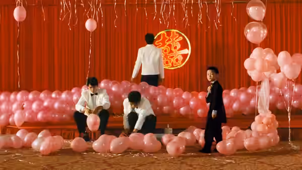 Yang-Yang stands among pink balloons as adults prepare a decorated celebration stage