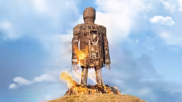 The wicker man stands ablaze on a grassy hill under blue skies