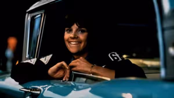 a smiling young woman rests her arms on the window of a blue car