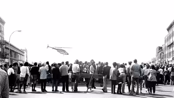 A large crowd gathers on a city street watching a helicopter overhead