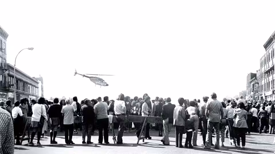 A large crowd gathers on a city street watching a helicopter overhead