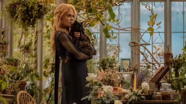 Nicole Kidman cradling a black cat inside a sunlit greenhouse filled with plants