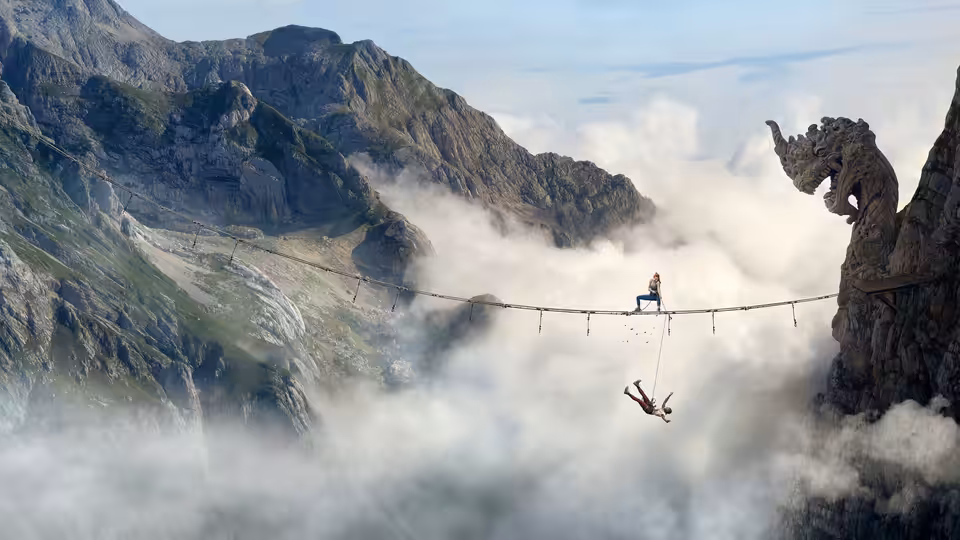 A climber sits on a highline while another hangs below over clouds