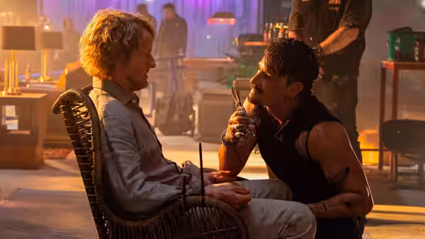 Ben (Owen Wilson) seated as Hank Malone (Alan Ritchson) holds a gun