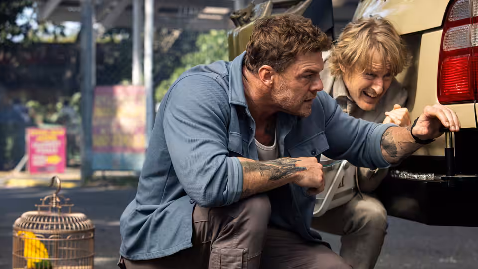 Hank Malone (Alan Ritchson) and Ben (Owen Wilson) crouching by a car