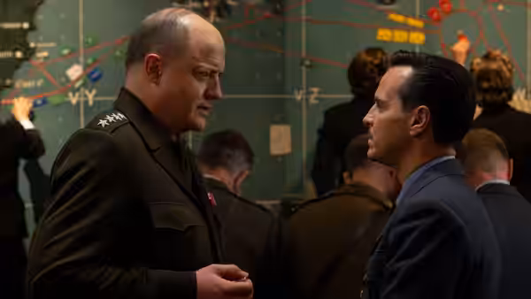Military officer speaks with man in suit before wall of maps