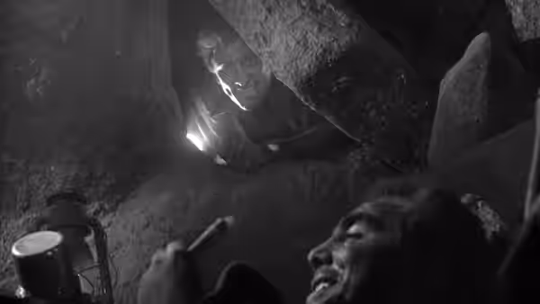 A man shines a flashlight down into a dark cave toward another person below