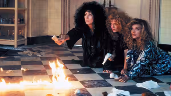 Alexandra Medford (Cher), Jane Spofford (Susan Sarandon) and Sukie Ridgemont (Michelle Pfeiffer) crouch beside flames on a tiled floor