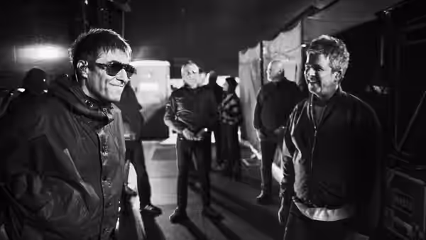 Two men talk face to face in a busy backstage area under lights