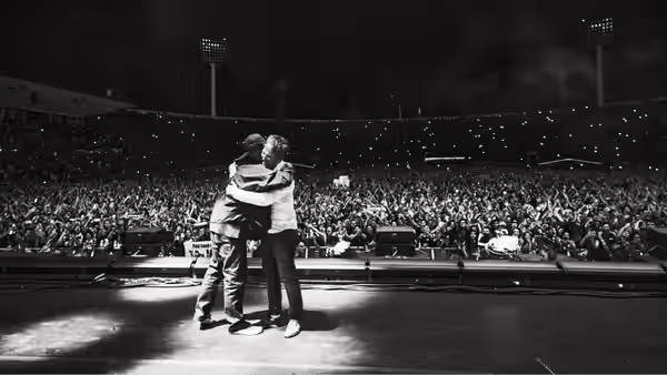 Two musicians hug tightly on stage in front of a packed stadium crowd