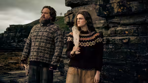 a bearded man and a woman in patterned sweaters stand on rocky cliffs
