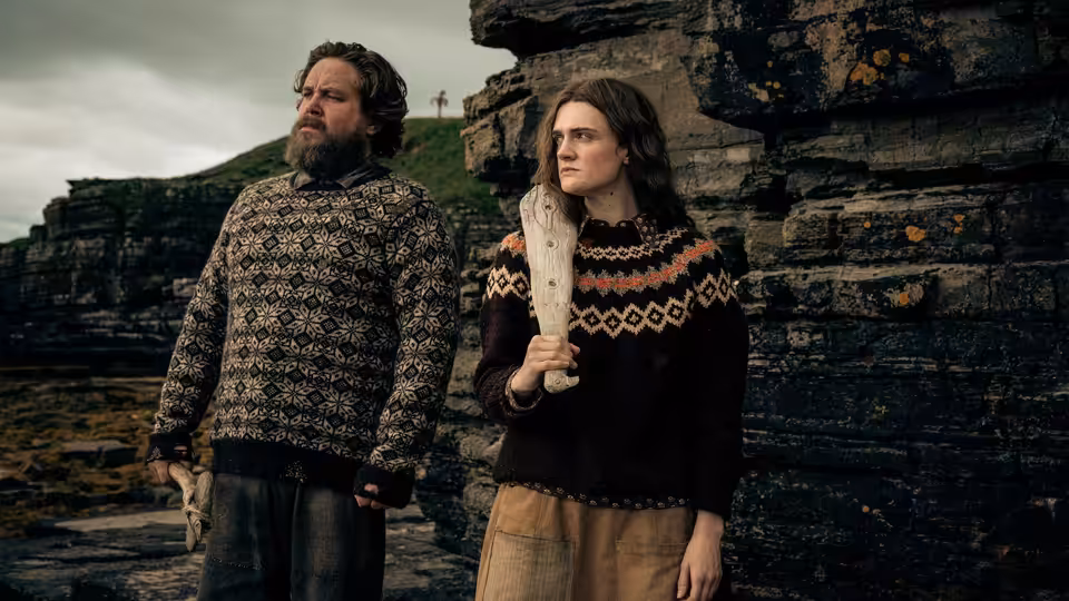 a bearded man and a woman in patterned sweaters stand on rocky cliffs