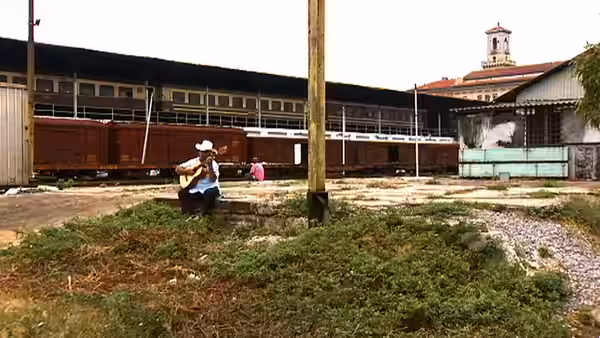 Man with guitar sitting on a wall beside train tracks and old buildings