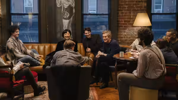Gloria (Greta Lee) and Ed Saxberger (Willem Dafoe) among a group in a lounge