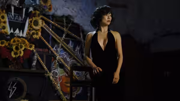 Gloria (Greta Lee) standing on a stage beside flowers and props